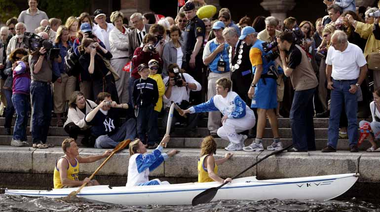 En kanot med tre kanotister vid kajen vid Stockholms slott, med Agneta Andersson i mitten, som får en fackla tänd av finansborgarrådet Annika Billström. 