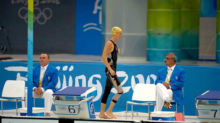 En kvinna i svart baddräkt och gul badmössa går förbi startpallarna vid en simbassäng. 