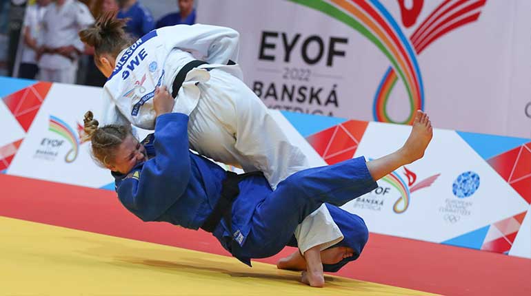 Två judokas i kamp, den i vita dräkten fäller den blåa i en benkrok. 