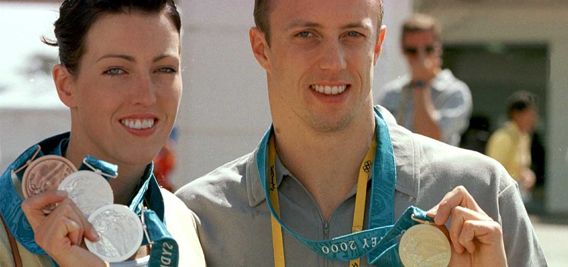 En kvinna och en man som håller armarna om varandras axlar och visar upp tre respektive en medaljer som hänger kring deras halsar