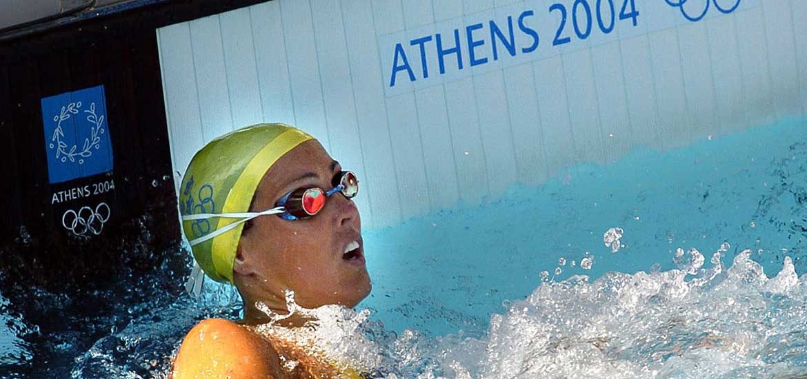 En kvinna i badmössa och simglasögon vid kortsidan i en simbassäng med logon för OS i Aten på kaklet
