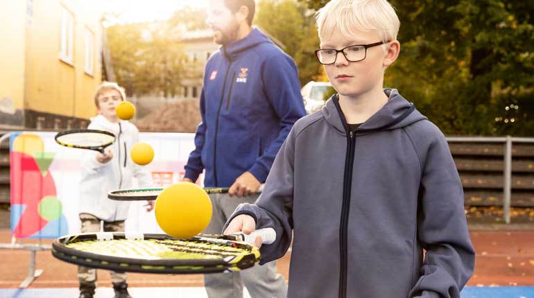Barn balanserar tennisbollar på tennisracket.