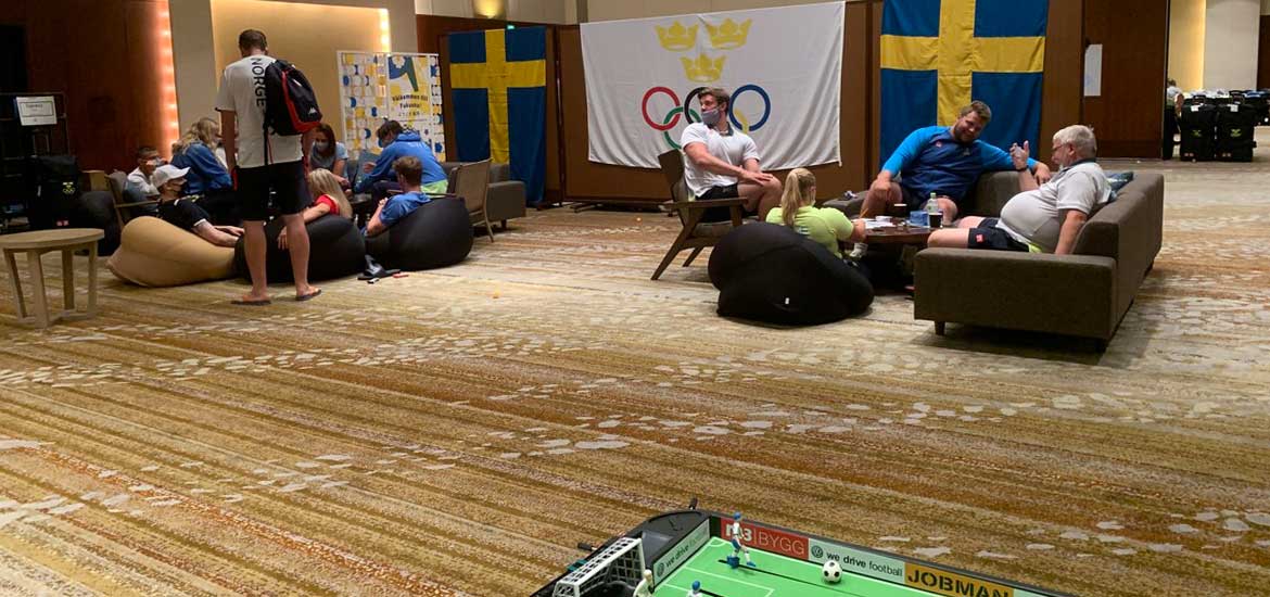 Personer sitter och pratar i en hotelllobbys soffor. Svenska flaggor i bakgrunden och ett fotbollsspel i förgrunden.