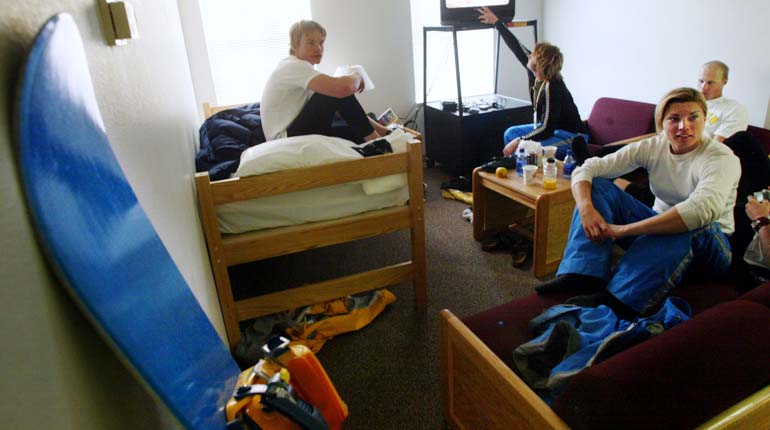 Tre personer sitter på sängarna i ett rum med våningssängar och en tv på en hylla. En snowboard står i förgrunden av bilden. 