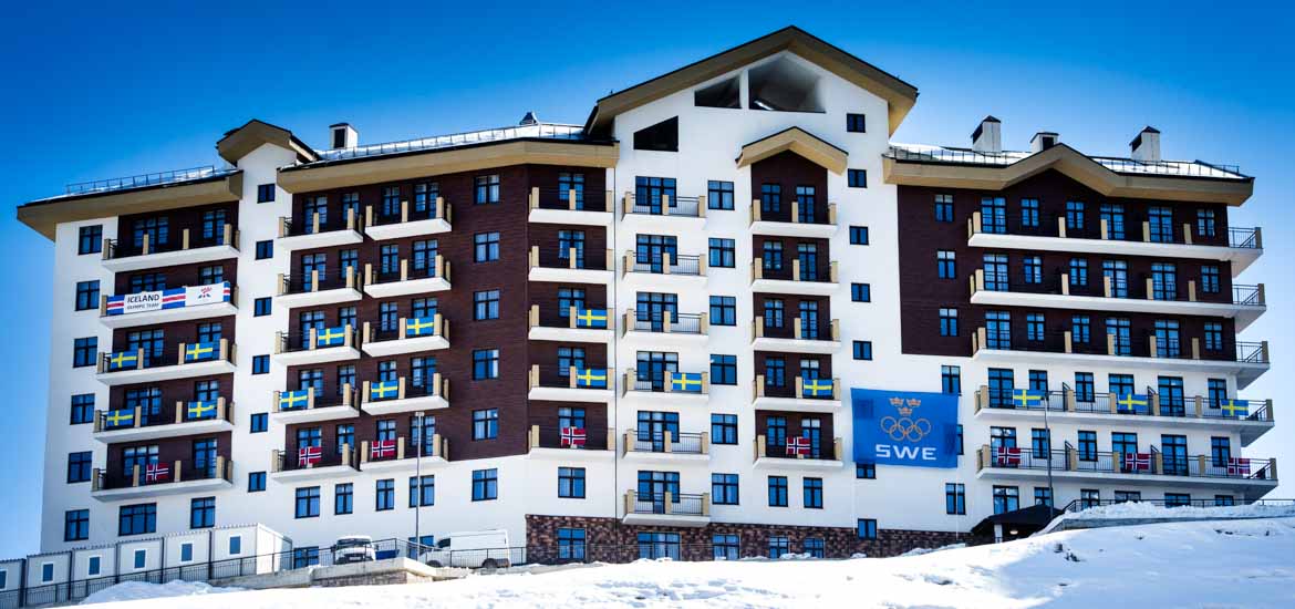 Ett stort lägenhetshus, sju våningar högt, med svenska och andra nationers flaggor längs med balkongerna på de olika våningarna. 