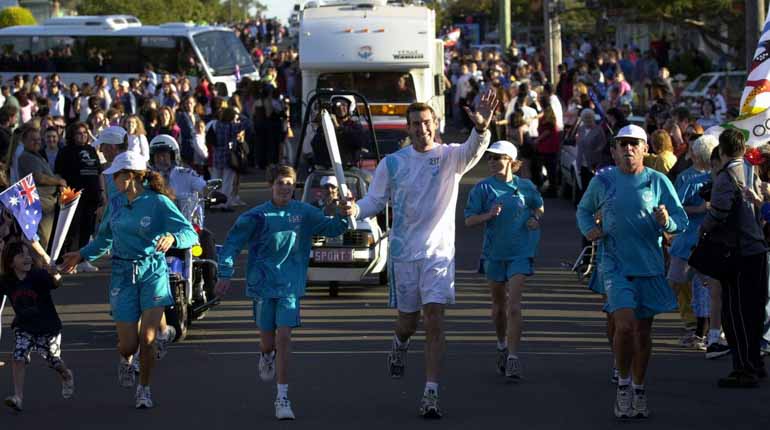 Man springer med den olympiska facklan omgiven av springande barn och massor av publik runtom. 