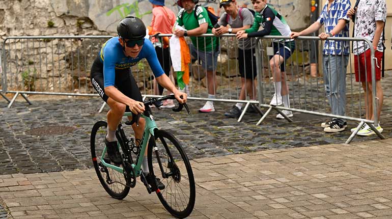 En cyklist i Sverigedräkt och en svartgrön biancicykel på kullerstensgator med publik i bakgrunden. Klotter på väggarna bakom. 