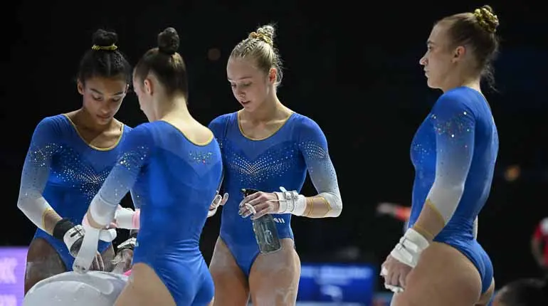 Fyra gymnaster i landslagsdräkter står och preparerar händerna med magnesium under en tävling. 